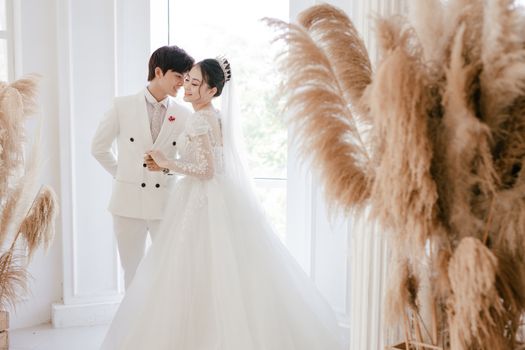 Happy gentle Asian young couple standing in wedding outfits near window in room decorated with feathers