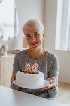 A senior woman with white hair holding a birthday cake with candles reading 56. Indoors setting.