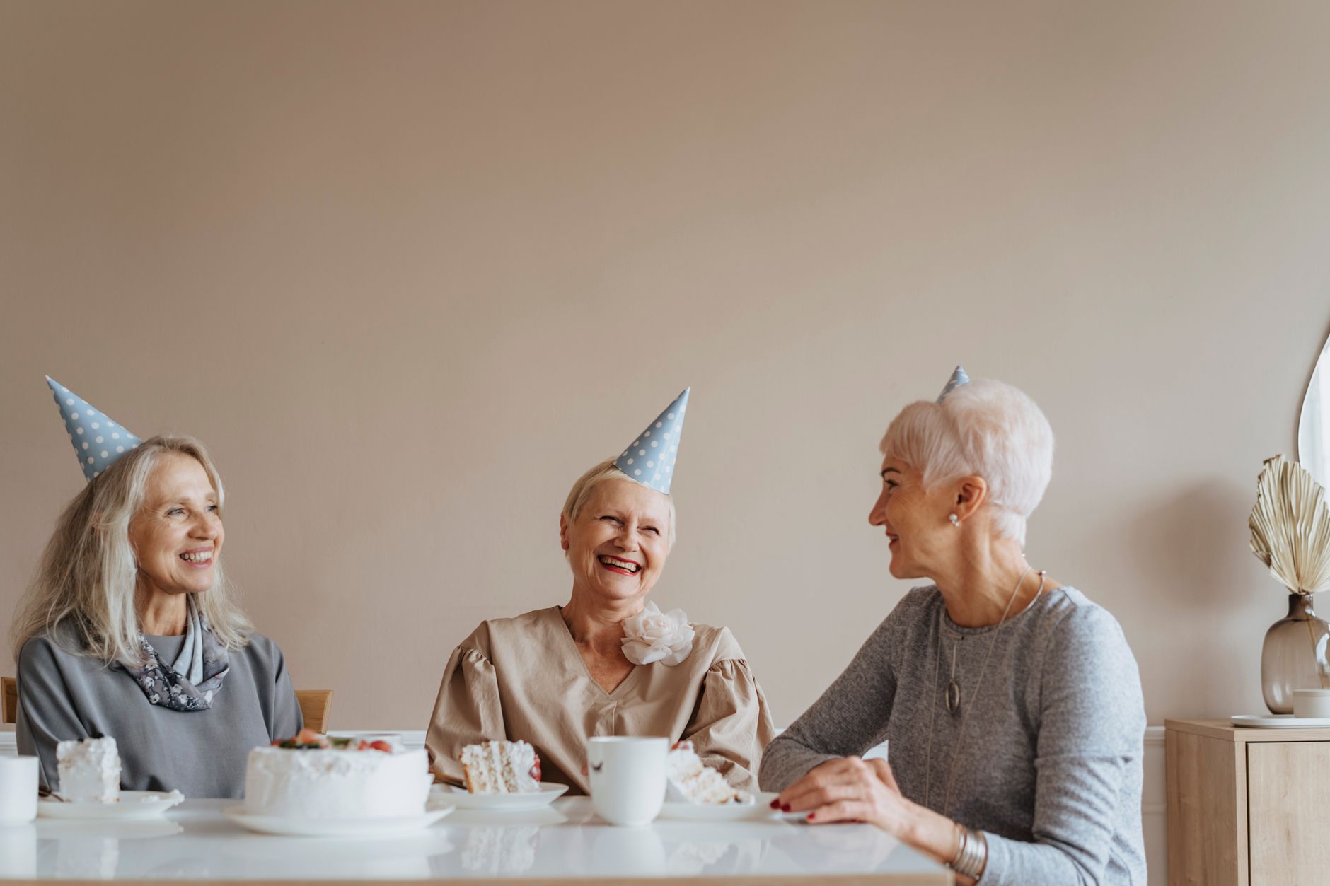 Three senior women enjoy a cozy indoor birthday celebration with party hats and smiles.