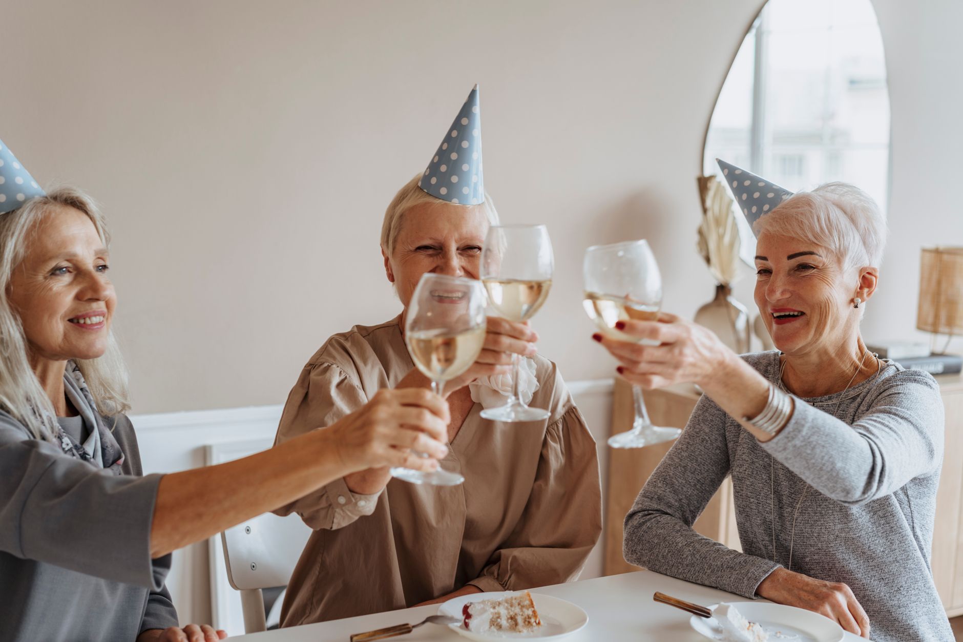 Three elderly women in party hats enjoying a birthday celebration indoors with drinks.