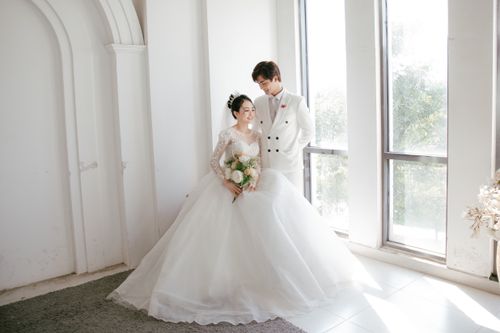 Free Smiling Asian bride with bouquet of flowers near groom in white suit standing in white studio in sunlight Stock Photo