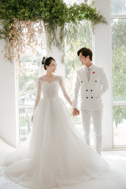 Full body of young Asian couple wearing white wedding outfits holding hands and smiling to each other