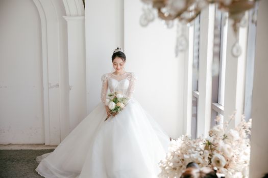 Smiling Asian bride in wedding dress with bunch of flowers standing near window and looking down