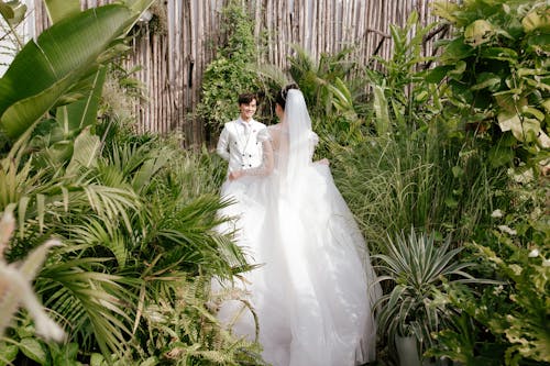 Bride in white wedding fluffy dress walking towards smiling Asian groom in green garden