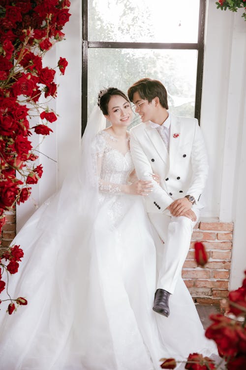 Cheerful Asian couple wearing white clothes sitting near window near red flowers at wedding day