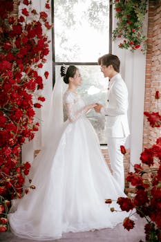 Newlywed couple holding hands, surrounded by red flowers indoors.