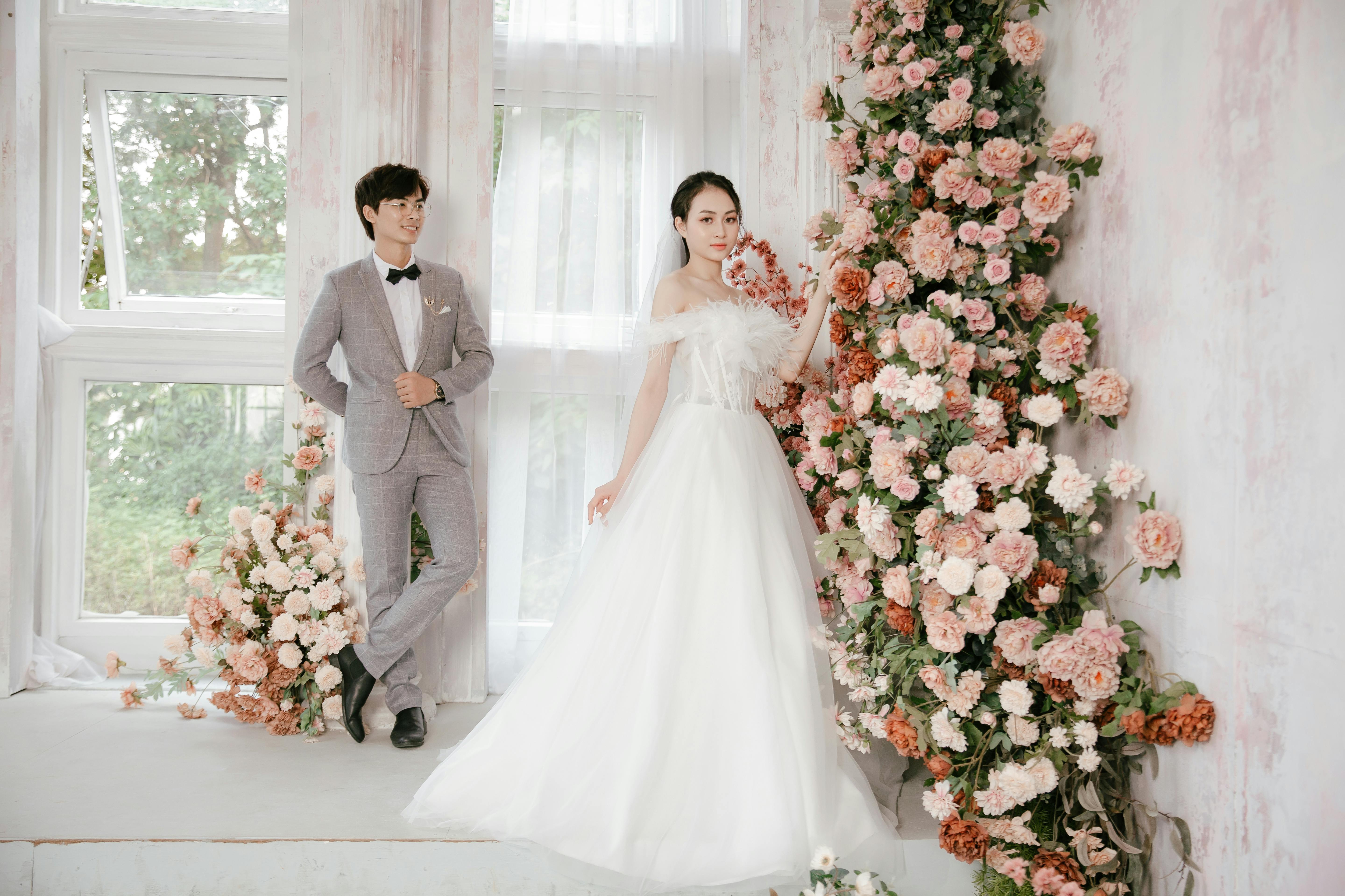 Beautiful Asian couple on their wedding day surrounded by pink and white floral arrangements indoors.