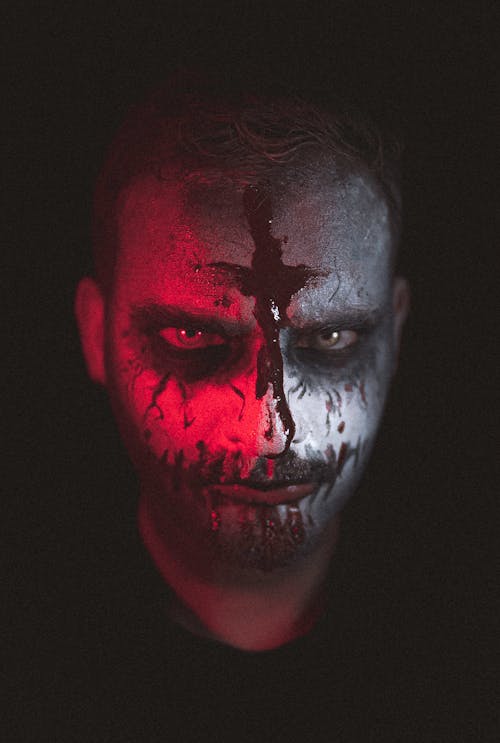 Spooky man with white face and black painted cross looking at camera in dark