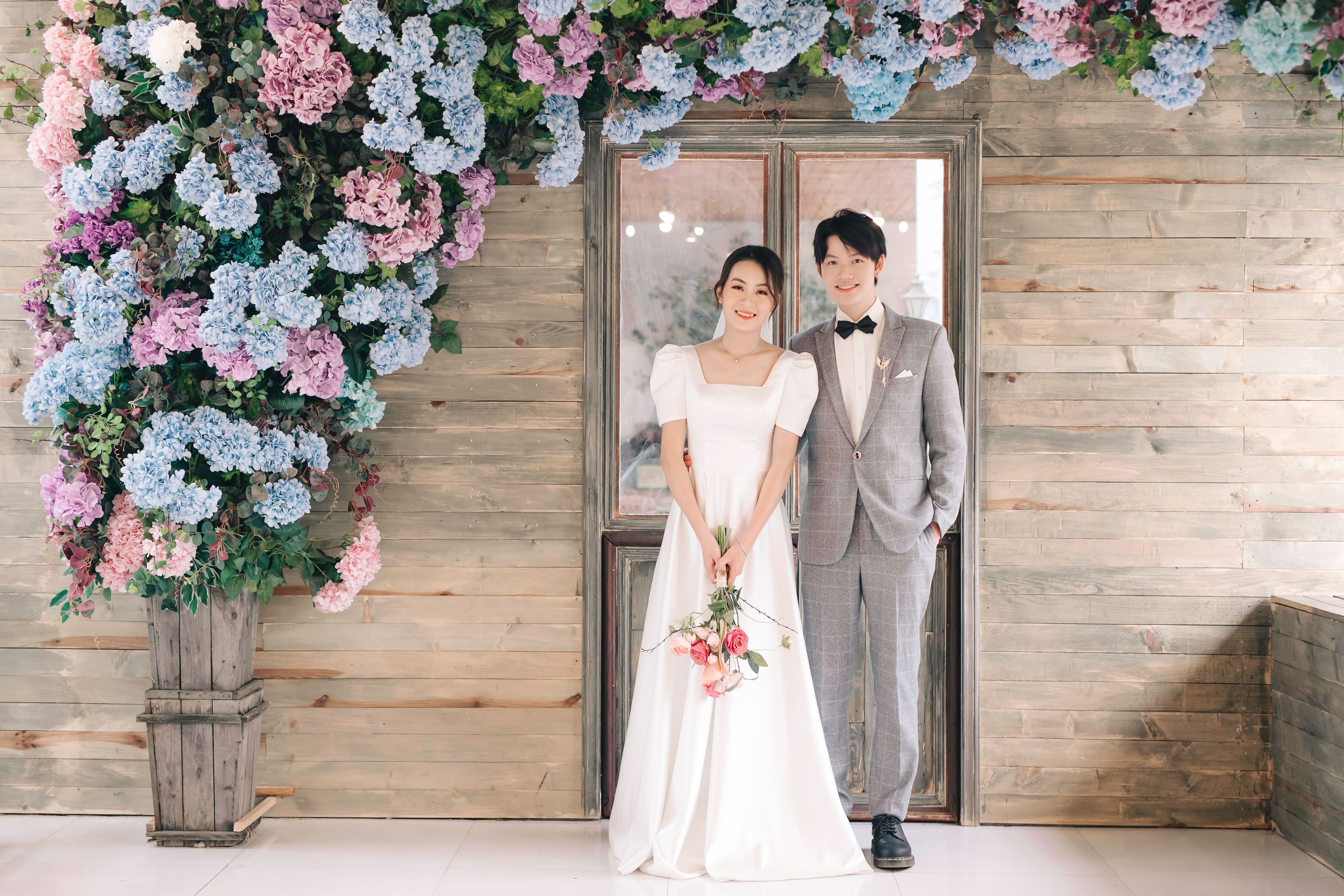 Elegant young couple posing in wedding attire against a floral backdrop indoors.