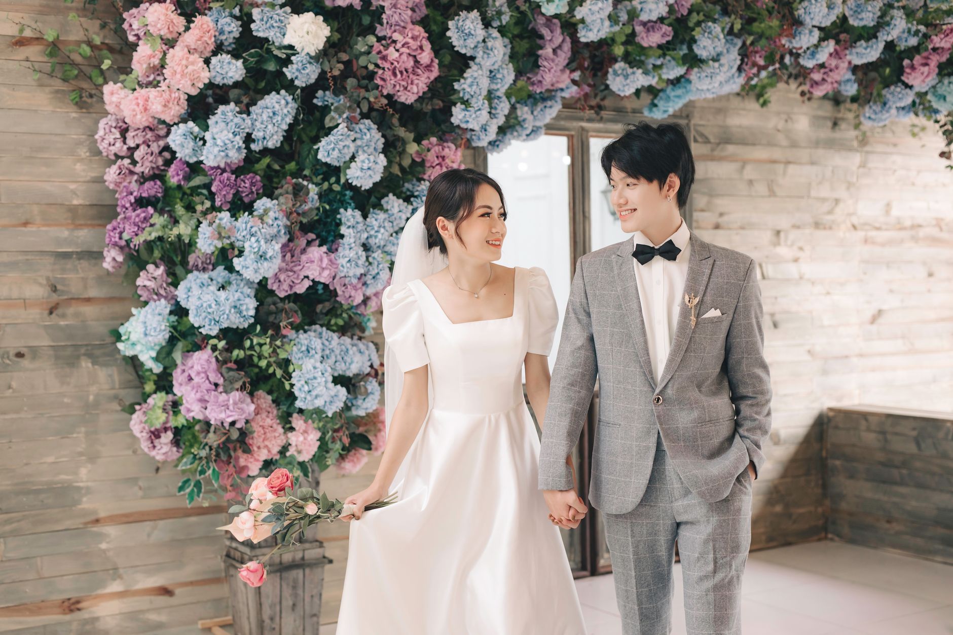 Romantic wedding photo of a couple holding hands surrounded by vibrant floral decor indoors.