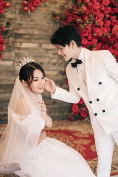Smiling Asian bride and groom in elegant attire surrounded by red roses on their wedding day.