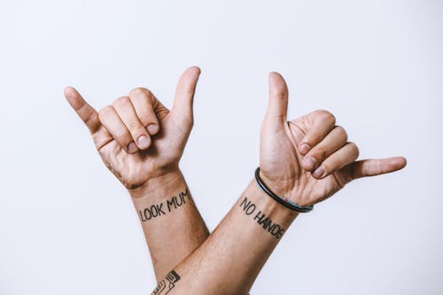 Crop anonymous male hands with creative look mum and no hands tattoo inscriptions showing shaka sign with fingers against white background