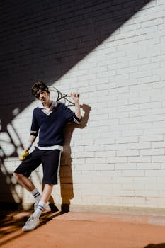 Stylish young man posing with tennis gear against a brick wall in sunlight.