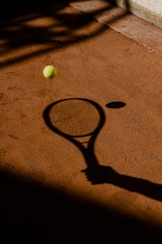 A tennis racket shadow and tennis ball on a clay court surface, capturing the essence of the sport.