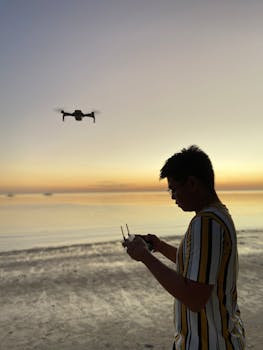 Silhouette of a man controlling a drone over a calm beach during sunset.