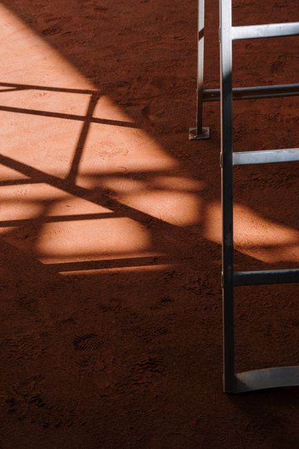 A ladder casts intricate shadows on a sunlit clay court, creating a geometric pattern.