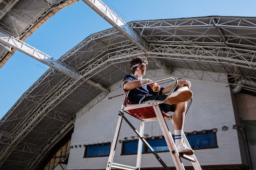 A young male referee sitting on a chair in a modern stadium, overseeing a tennis game. Bright daylight enhances clarity.