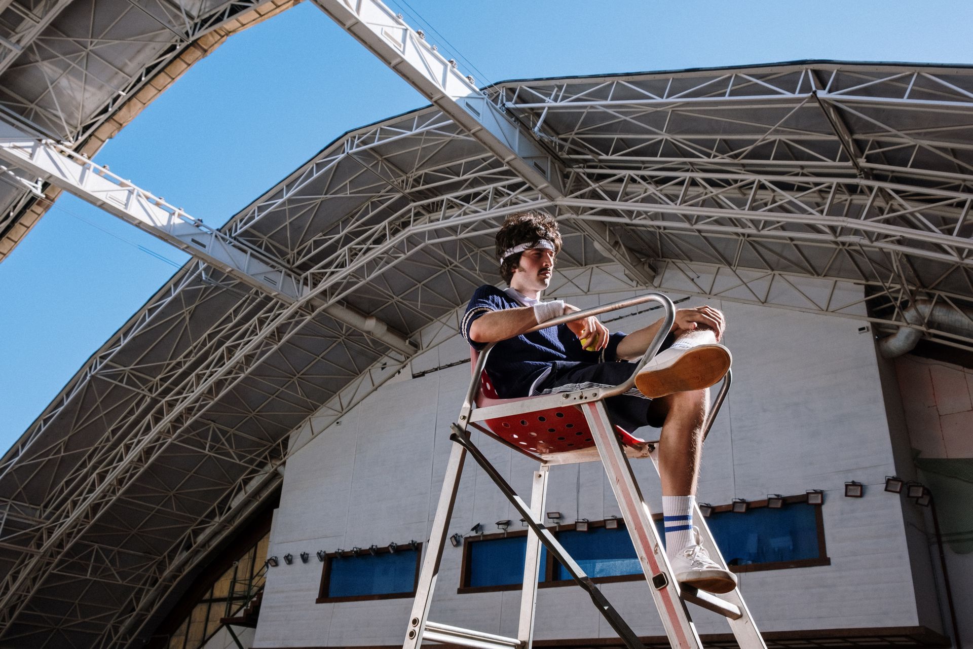 A young male referee sitting on a chair in a modern stadium, overseeing a tennis game. Bright daylight enhances clarity.