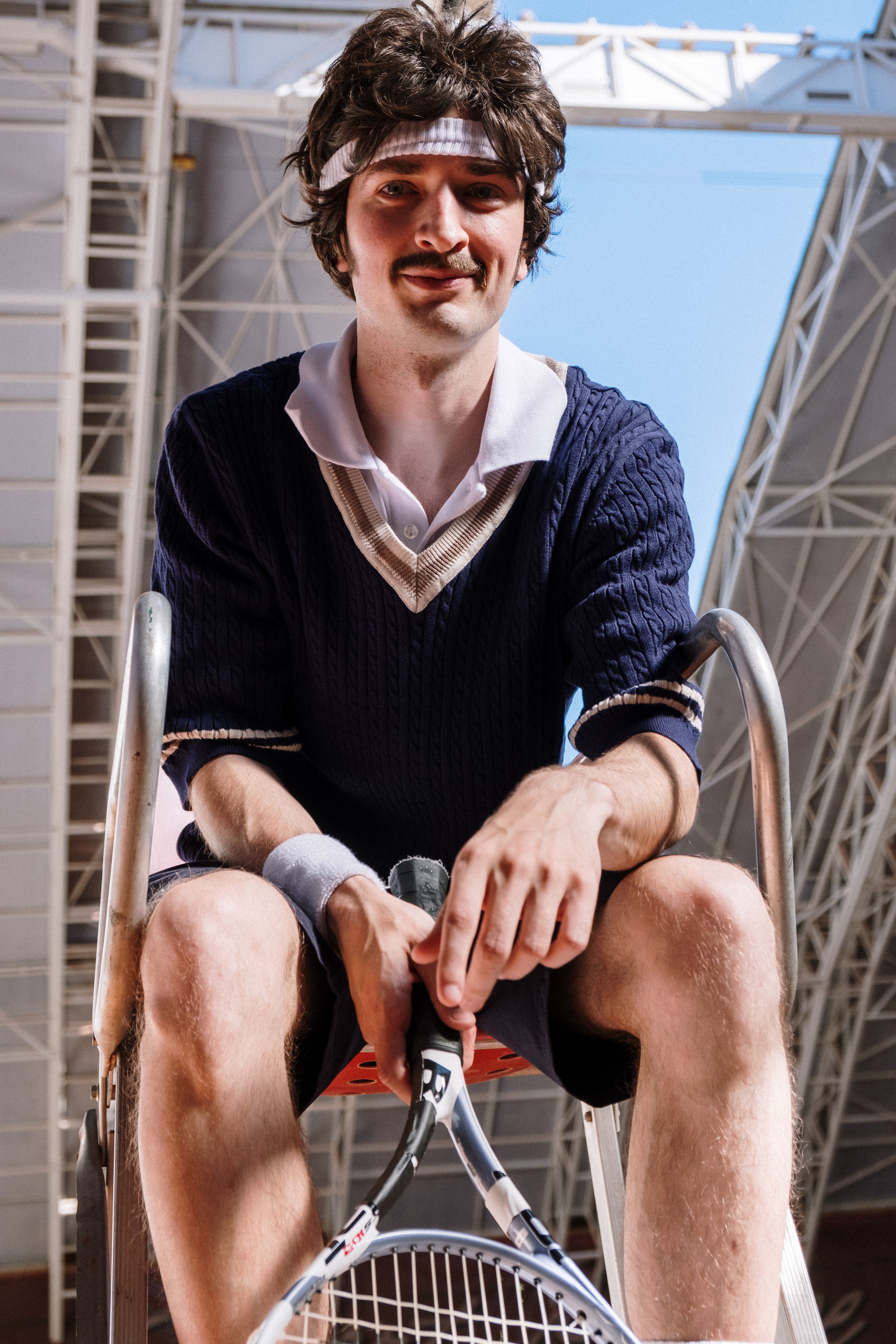 一名身穿70年代网球服的白人男子拿着球拍，坐在户外体育场里