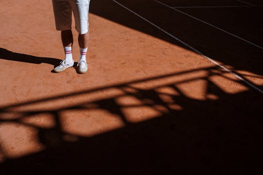 An athlete's legs and sneakers casting a shadow on a sunlit clay tennis court.