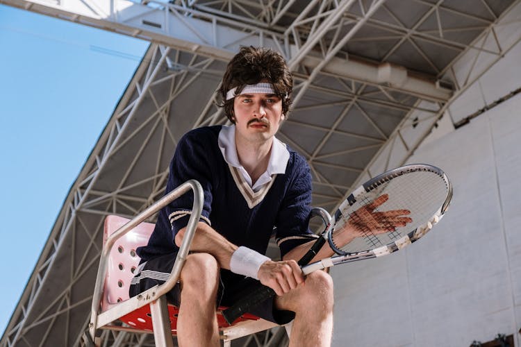 Man Sitting On Tennis Umpire High Chair Holding A Tennis Racket