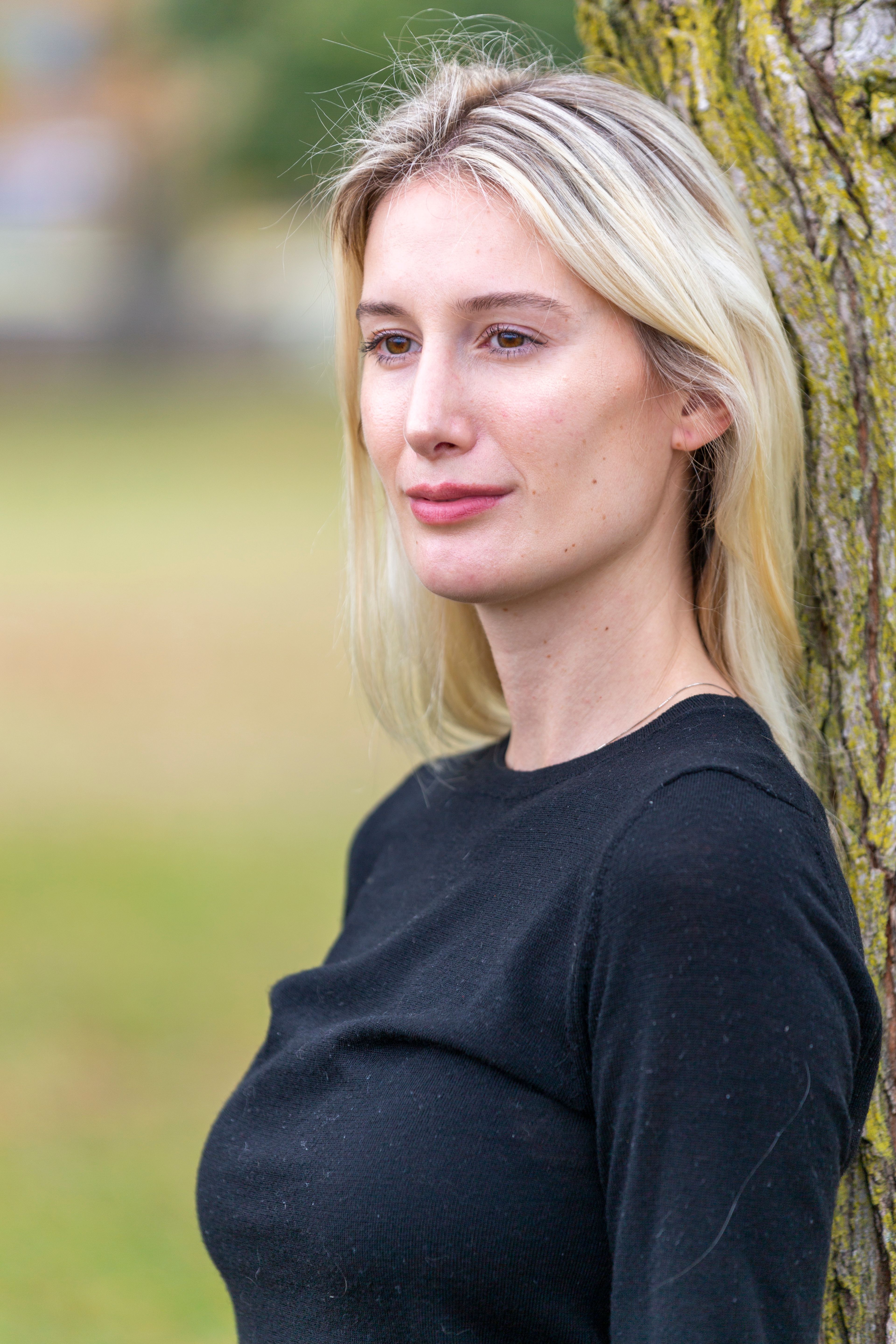 Slender young dreamy female with fair hair looking away near tree on blurred background of nature