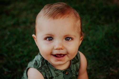 From above of cute baby in ornamental wear smiling while looking at camera on lawn