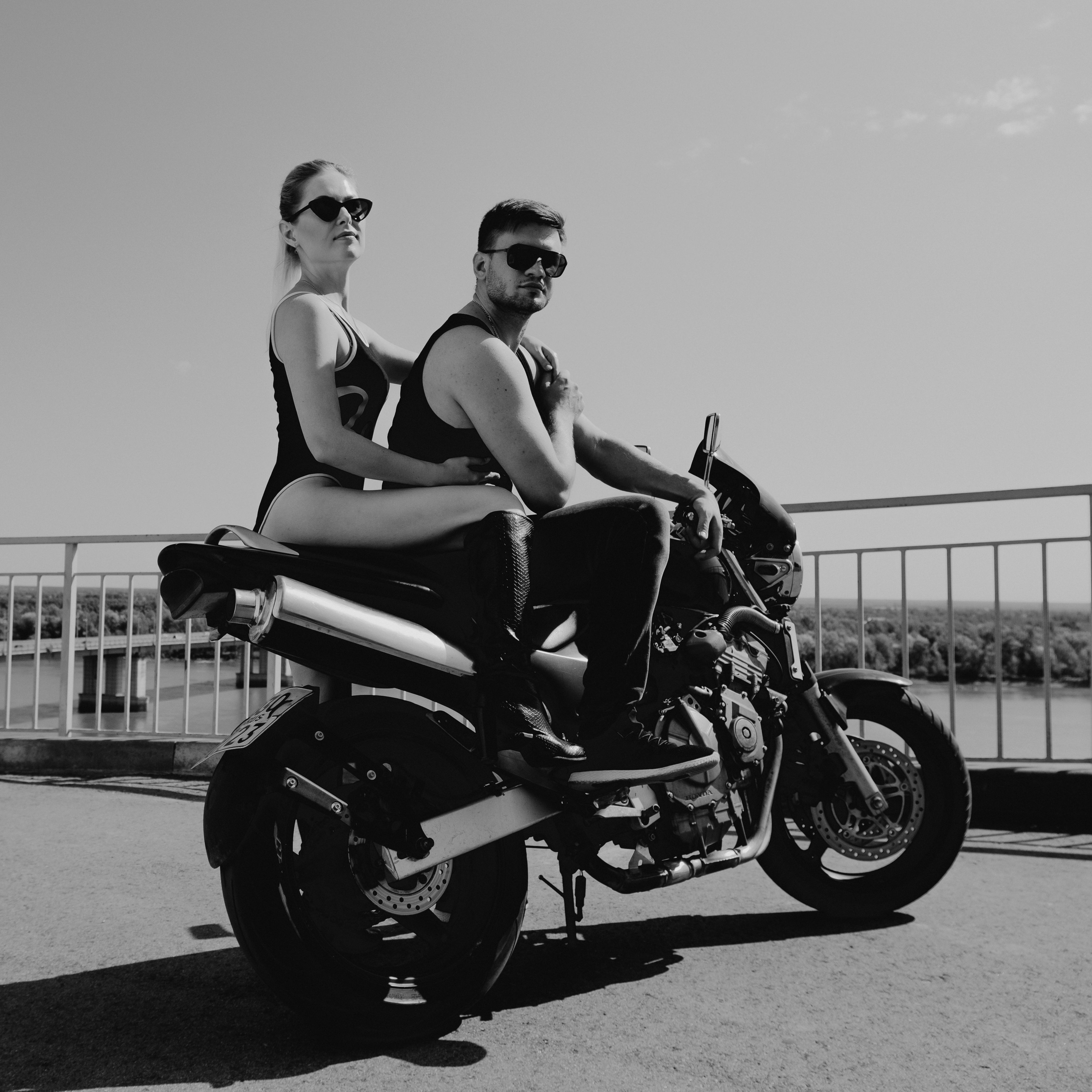 Black and white of confident young couple in sunglasses looking at camera while hugging and sitting on motorcycle on asphalt road near railing above river under cloudless sky in daytime