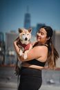 阳光明媚的日子里，一位微笑的女人抱着她的柴犬，对着城市天际线