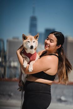 阳光明媚的日子里，一位微笑的女人抱着她的柴犬，对着城市天际线