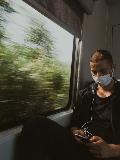 A man wearing a face mask and earphones uses his phone while traveling on a train, with blurred motion outside.
