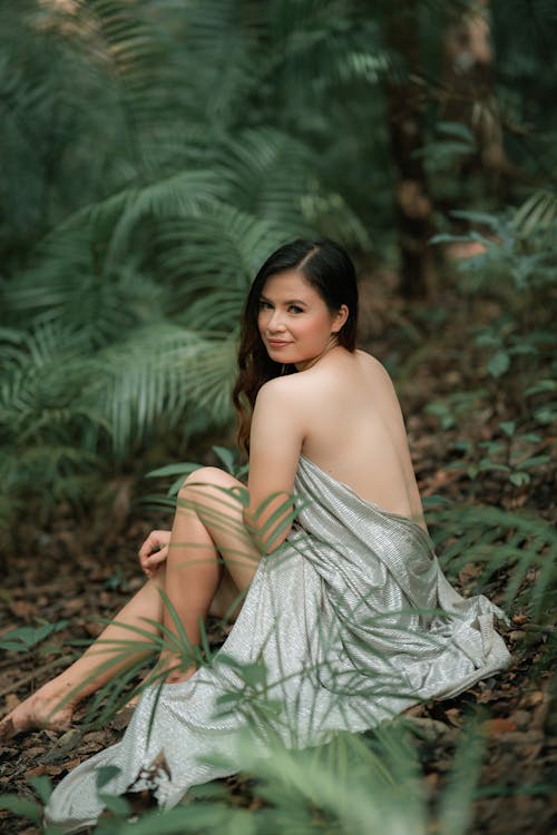 Back view of young sensual ethnic female wrapped in drapery with makeup looking at camera while resting among fern plants