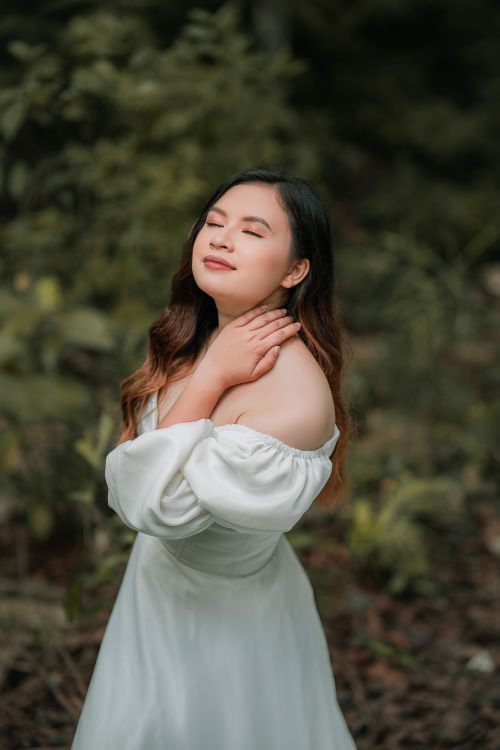 Free Side view of young mindful ethnic female with closed eyes in elegant apparel touching neck near bushes in park Stock Photo
