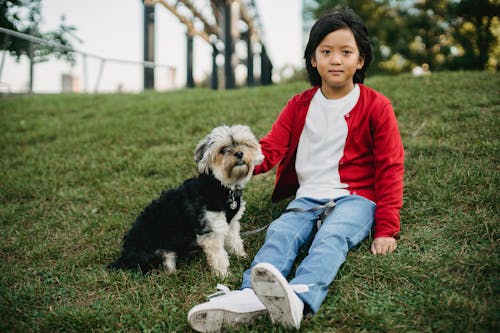 Charming ethnic kid resting on meadow with little dog while looking at camera in city