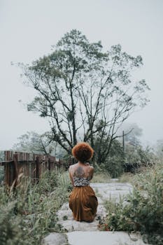 一名妇女独自坐在薄雾缭绕的小路上，小路四周环绕着郁郁葱葱的绿树，环境宁静而乡村