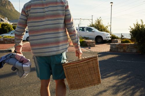Free A person carrying a woven picnic basket and blanket in a sunny outdoor parking area. Stock Photo