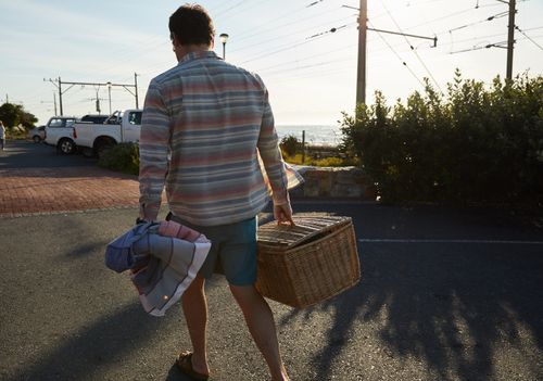 Free A man in a striped shirt carries a wicker basket and towel at a seaside parking area during the day. Stock Photo