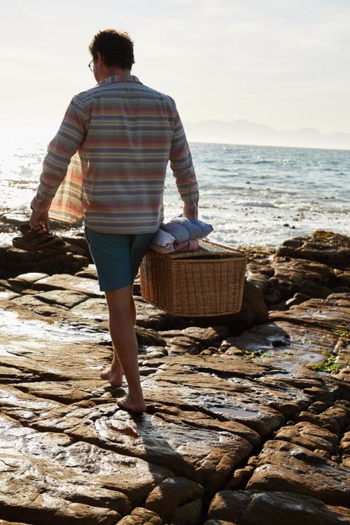 Free Man strolling barefoot on rocky coast carrying a wicker basket with summer essentials. Stock Photo