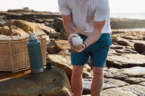 Free Man preparing coffee near a wicker basket on a rocky beach during a sunny day. Stock Photo