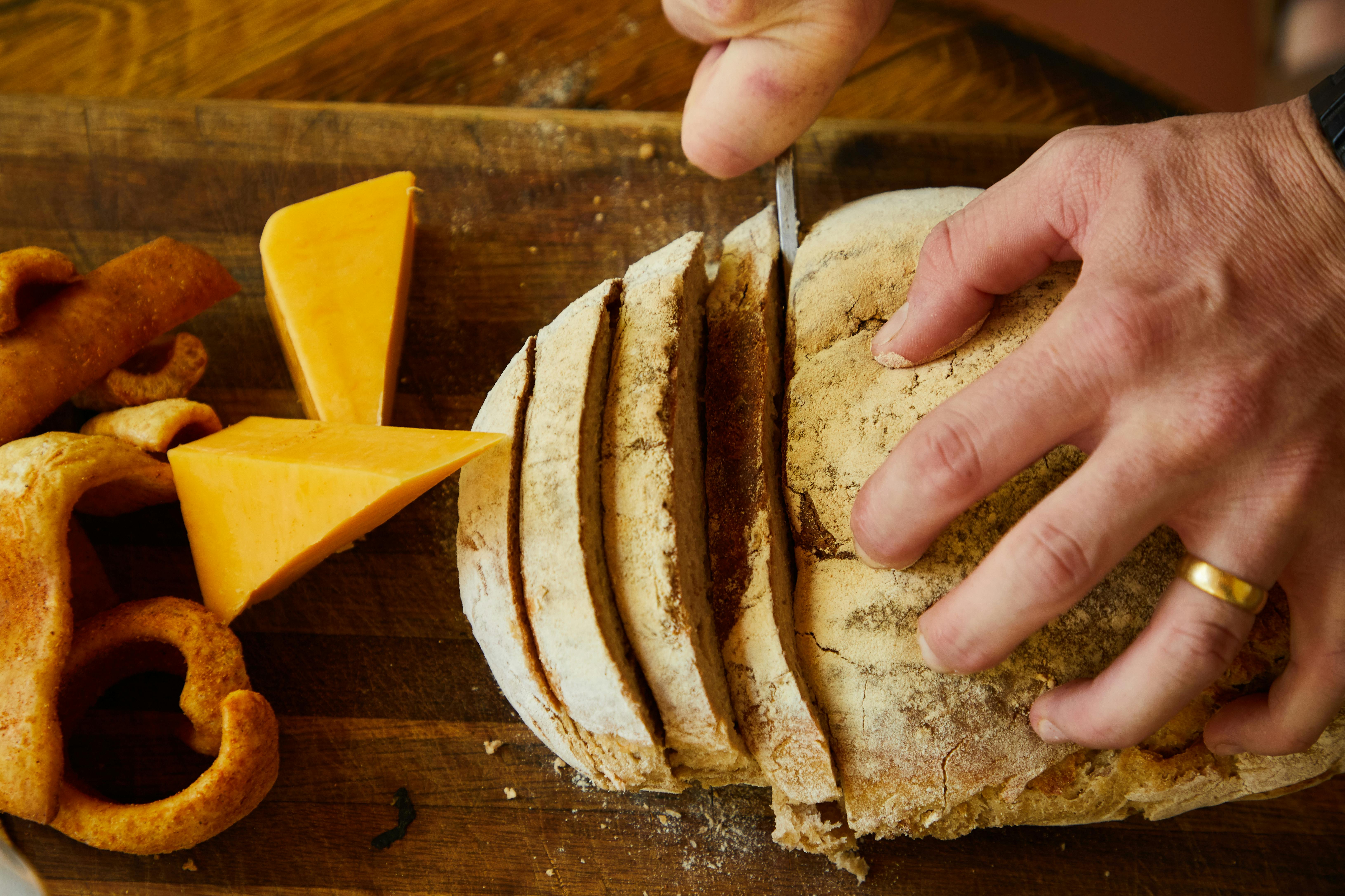 Close-up of homemade bread being sliced on a wooden board with cheese and snacks around. Ideal for a rustic culinary setting.