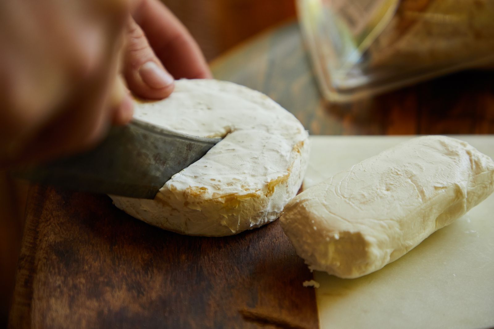 A small round of white homemade cheese being cut with a knife on a wooden board