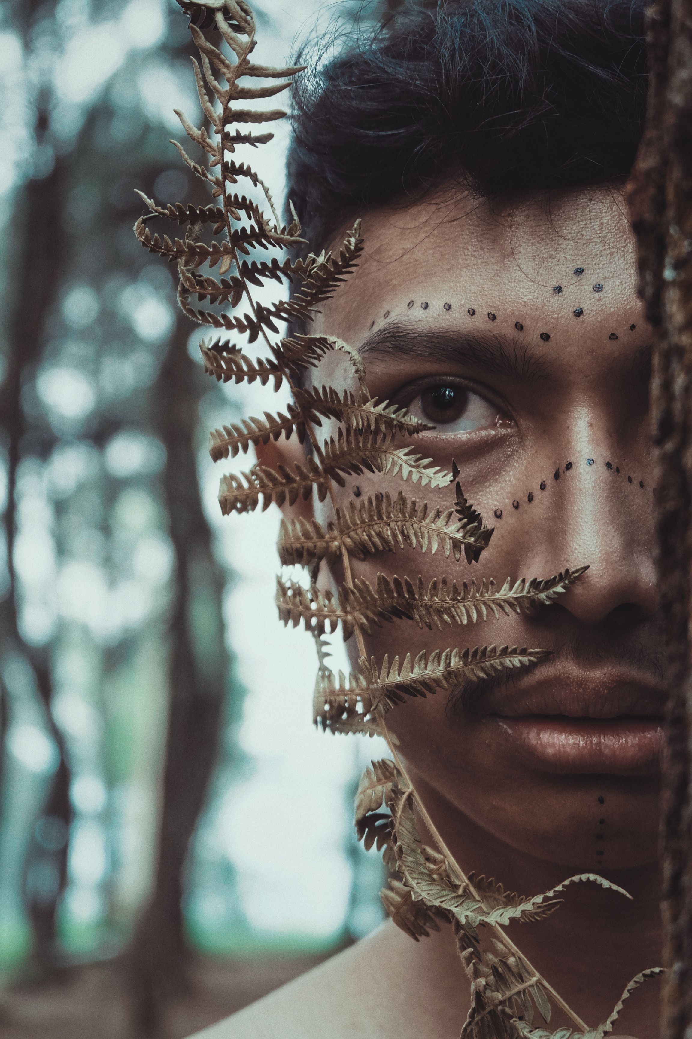 A man with artistic face paint partially hidden by a tree in a forest setting, conveying a natural and mysterious vibe.