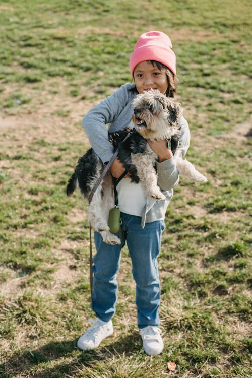 Content Asian boy carrying adorable purebred dog while standing on meadow and looking forward