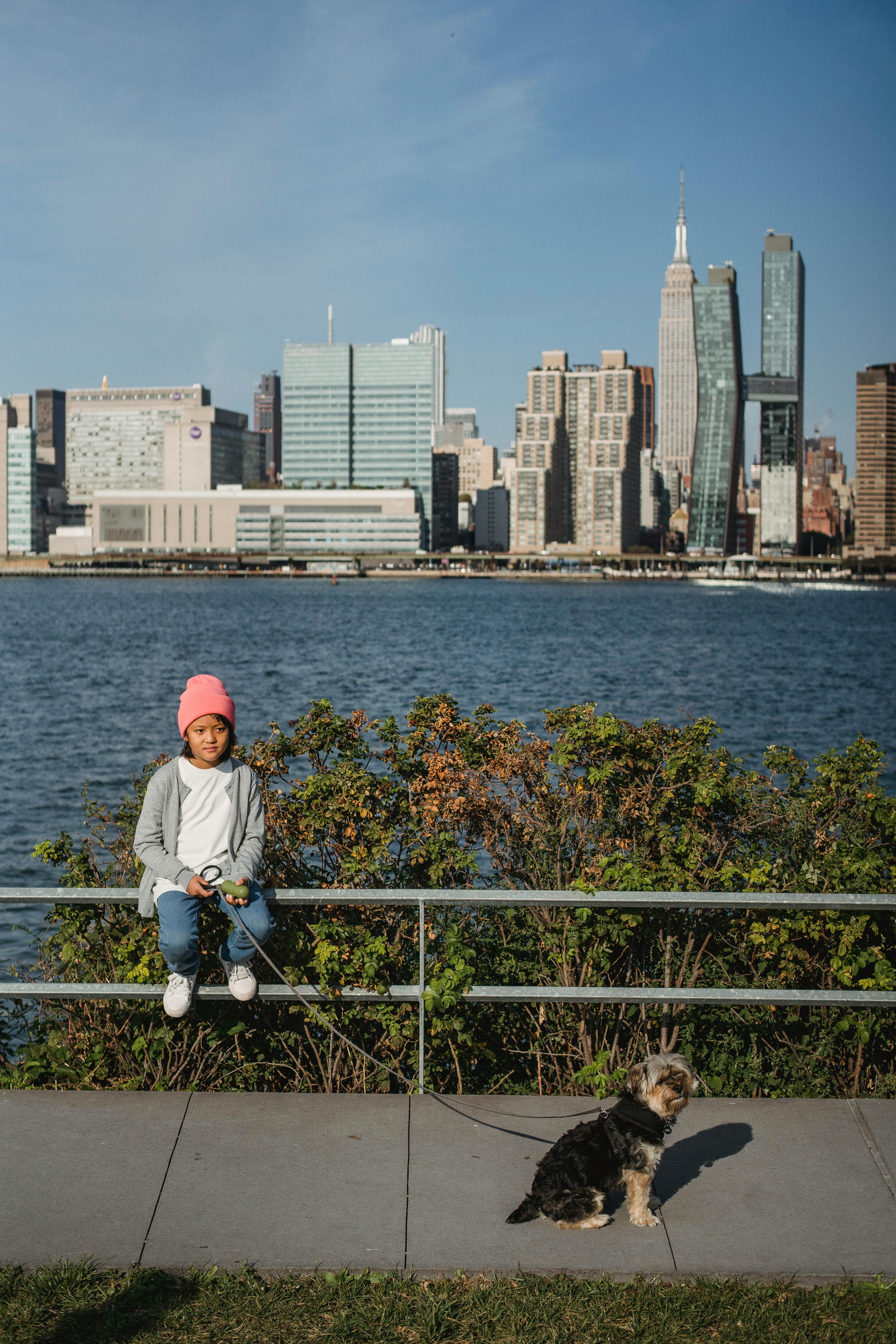 Full body of ethnic child sitting on metal fence and holding leash with puppy while spending time together