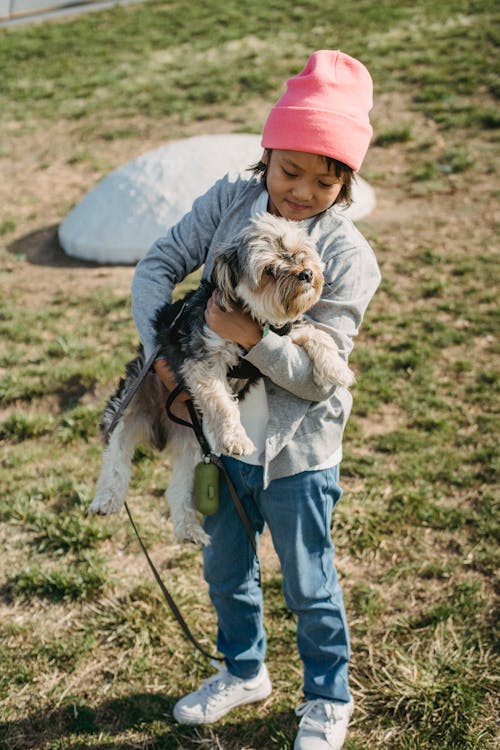 Full body of cheerful ethnic child in casual clothes standing on grass and embracing puppy