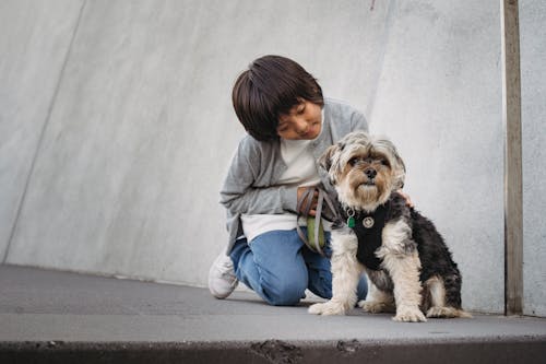 Cheerful ethnic child in casual clothes sitting on knees while stroking fluffy puppy on pavement