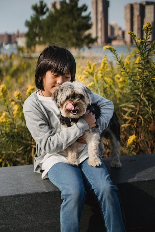 Free Ethnic child cuddling puppy while sitting on stone border in park with lush vegetation Stock Photo