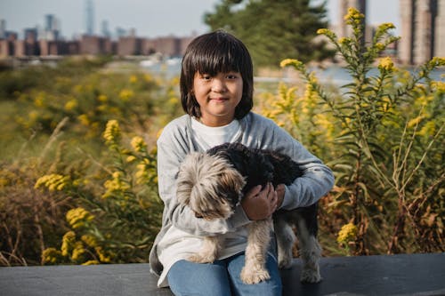 Happy little boy with modern haircut hugging fluffy dog while relaxing in park and looking at camera