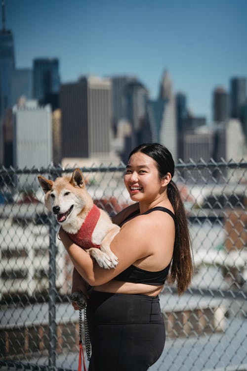 Side view of young ethnic female in sportswear standing near lattice fence and hugging cute puppy while looking at camera