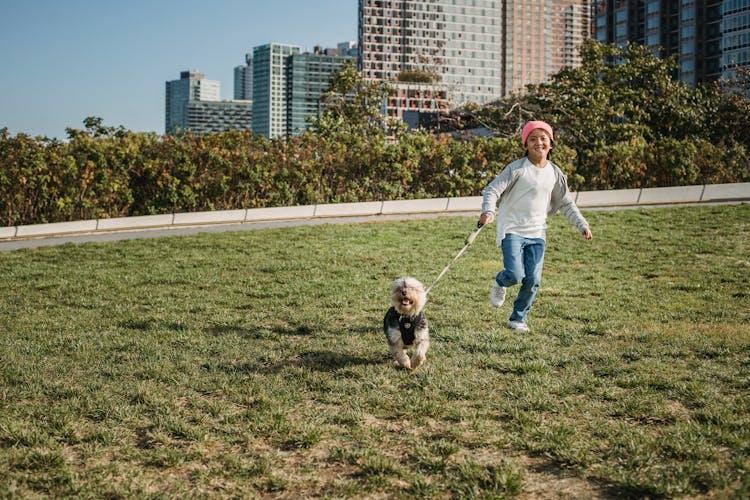 Happy Asian Boy Running With Dog On Leash On Meadow